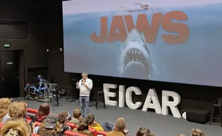 Damien Maric, micro en main, sur la scène de la salle de cinéma d’EICAR devant l’affiche des Dents