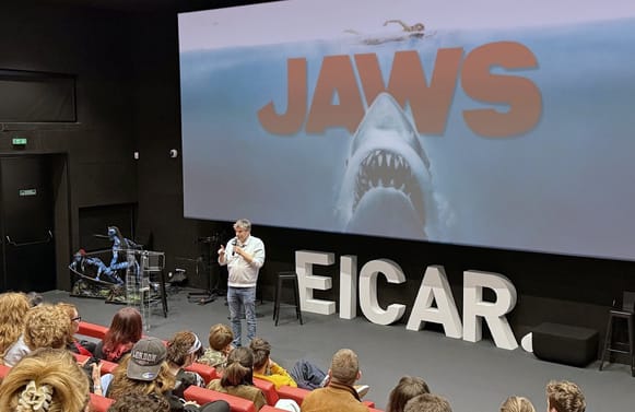 Damien Maric, micro en main, sur la scène de la salle de cinéma d’EICAR devant l’affiche des Dents